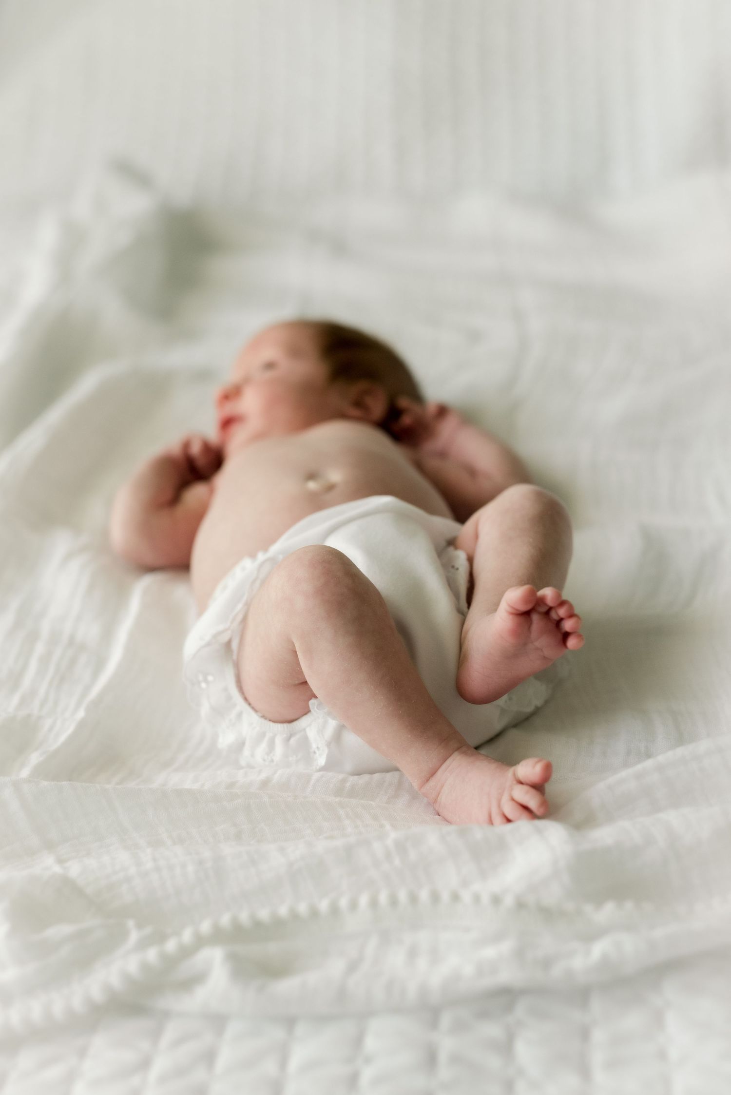 Newborn baby wearing white stretches out on cozy white bedding.