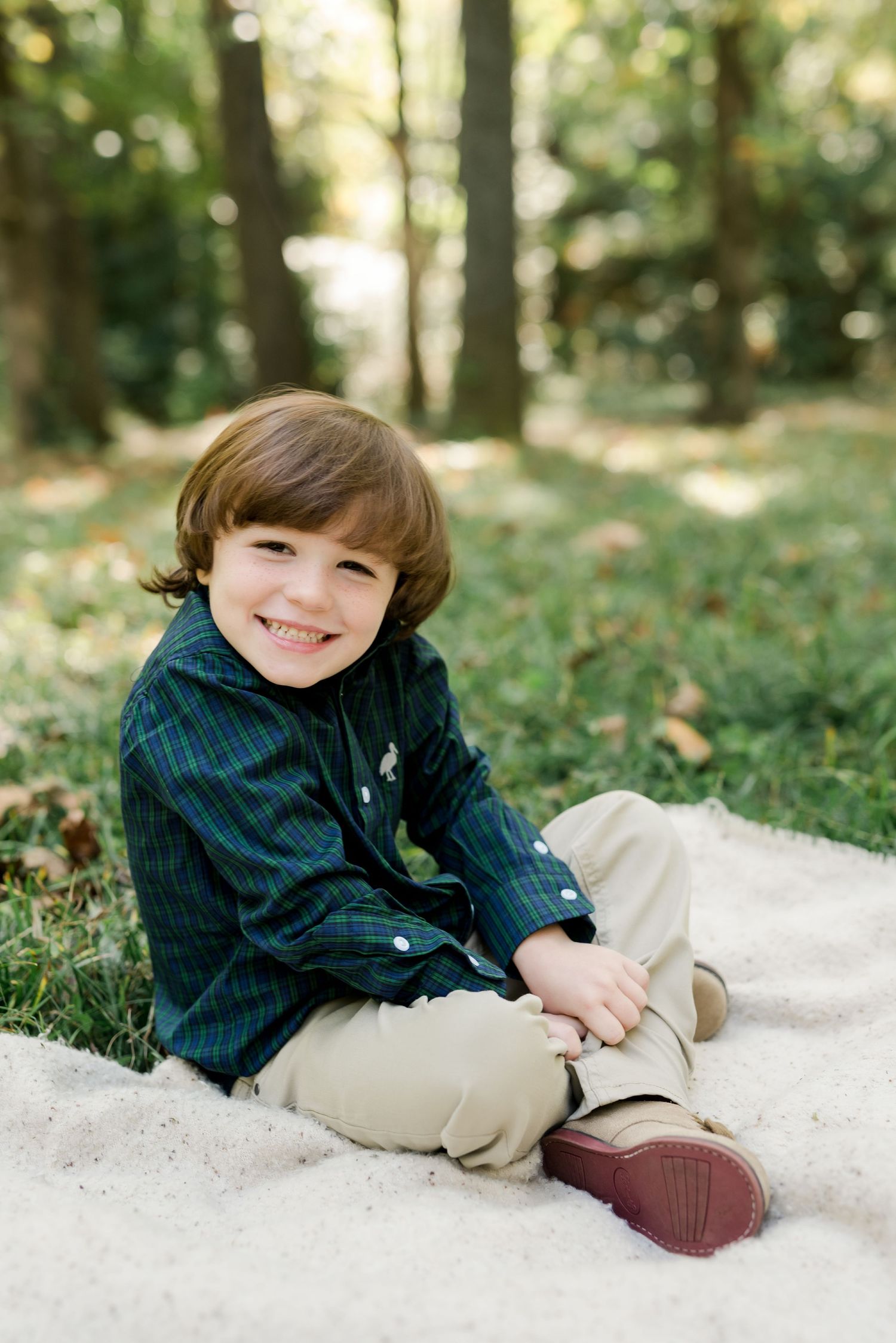 Child in striped button-down shirt and khaki pants sits on blanket in wooded setting.