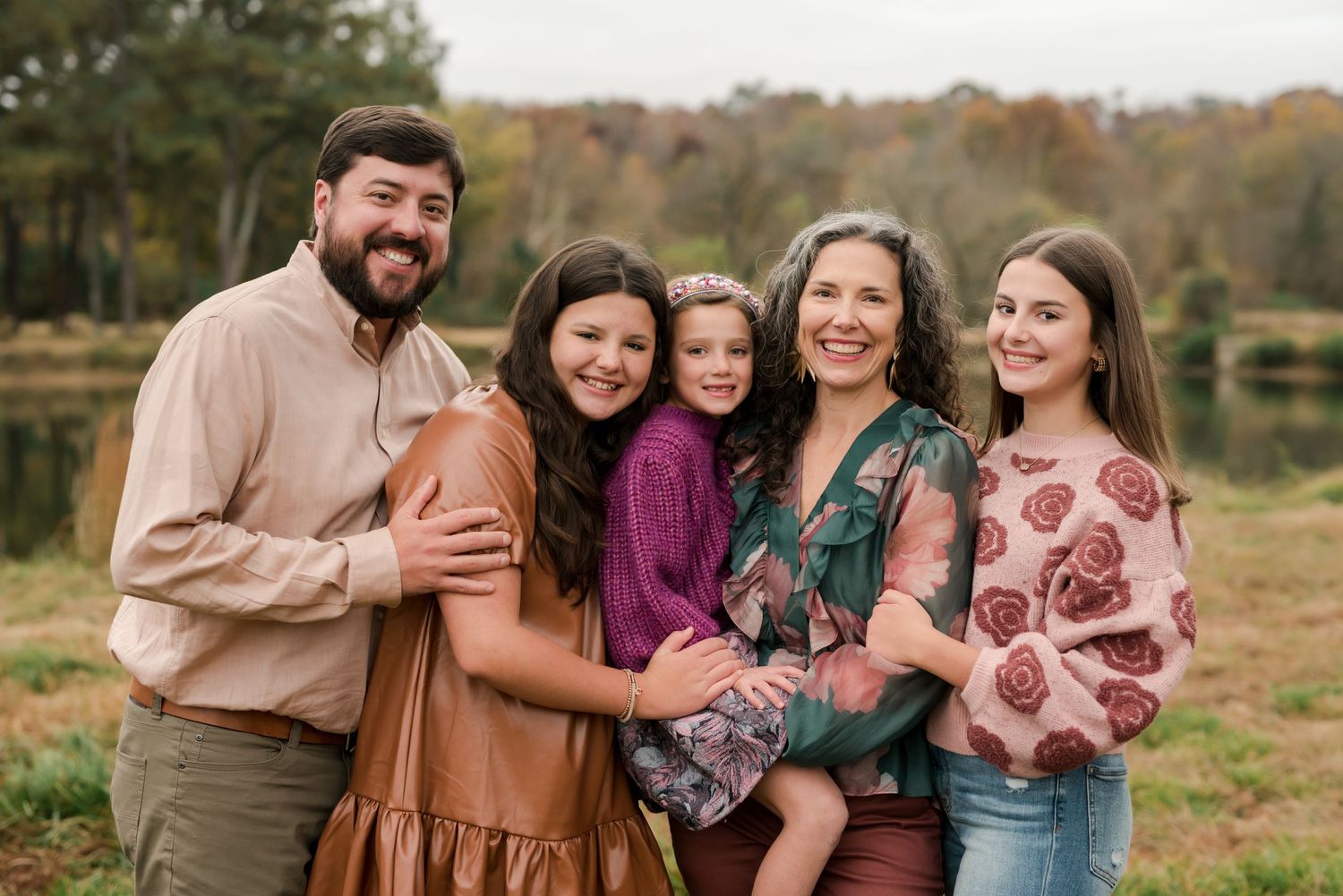 Candid family portrait of five people embracing outdoors with a lake and trees in the background.