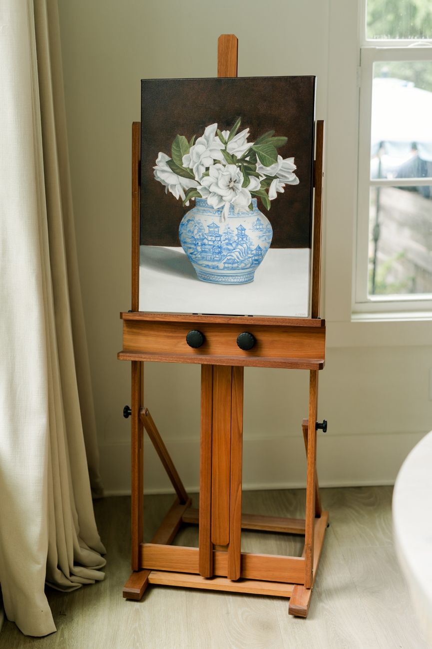 Wooden easel displaying framed artwork in bright interior room with white curtains.