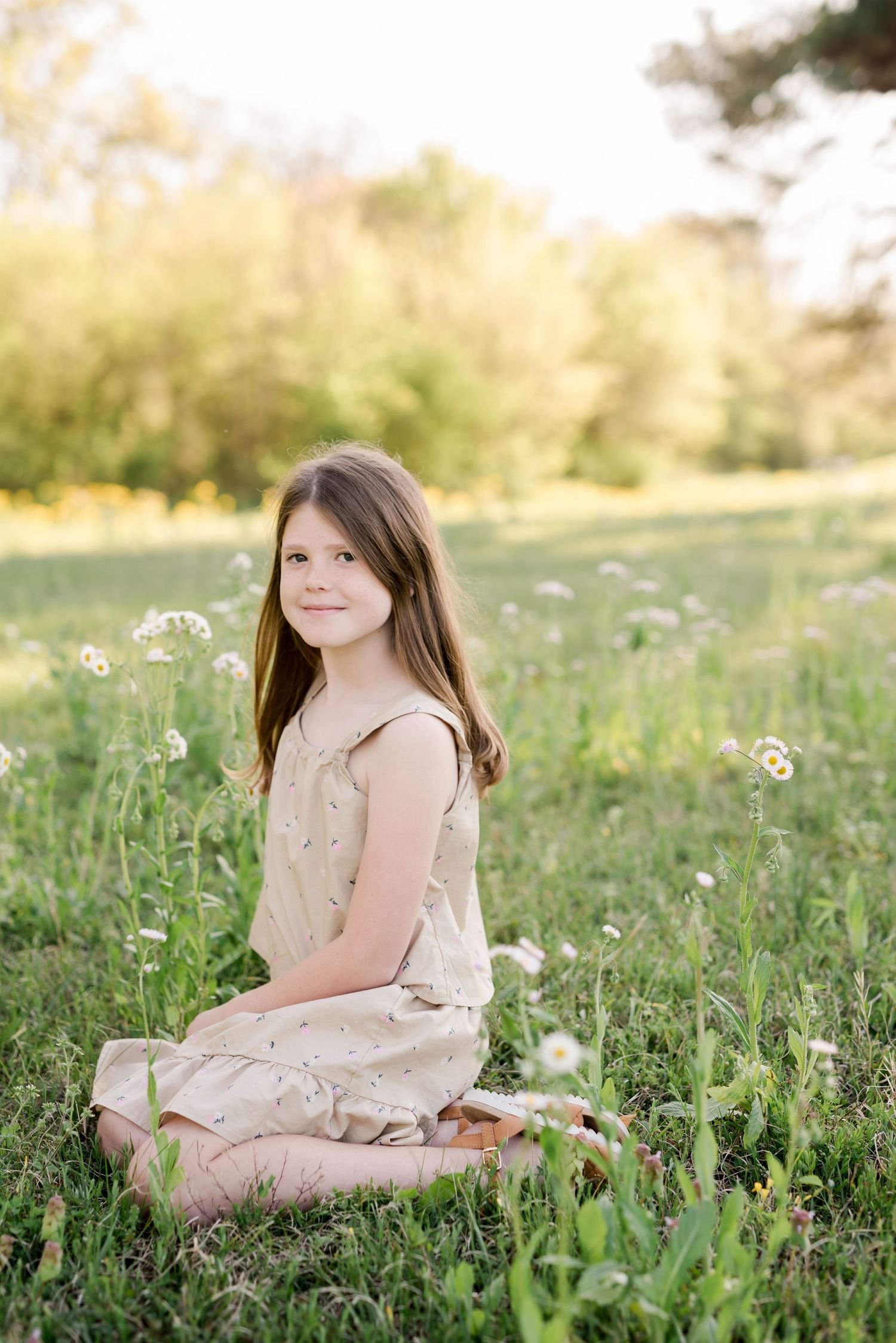 A child in a beige dress sits peacefully among wildflowers in a spring meadow.