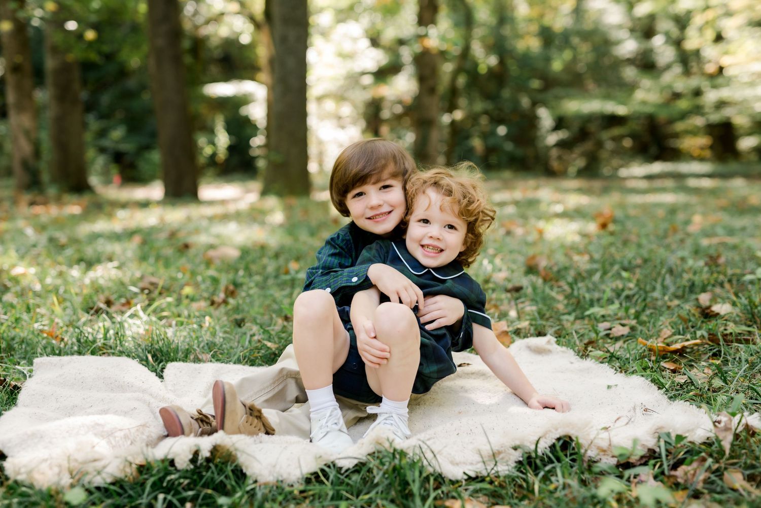 Two children sitting together on a white blanket in a sunlit wooded area.
