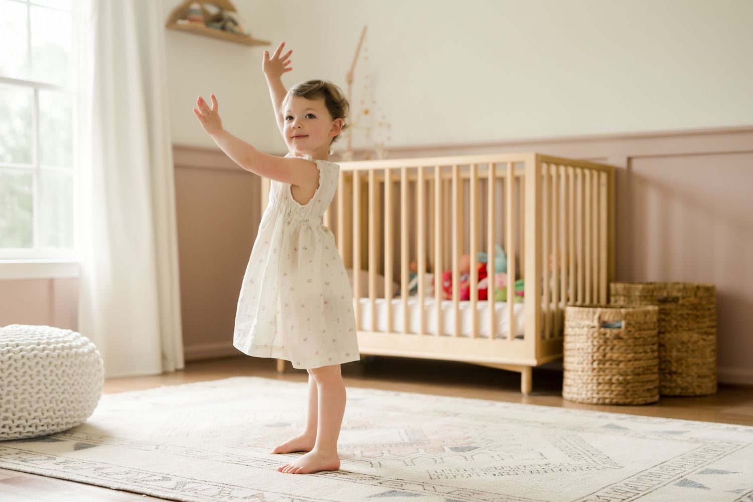 Young child in white dress playing and dancing in sunny nursery room with wooden crib and basket decor.