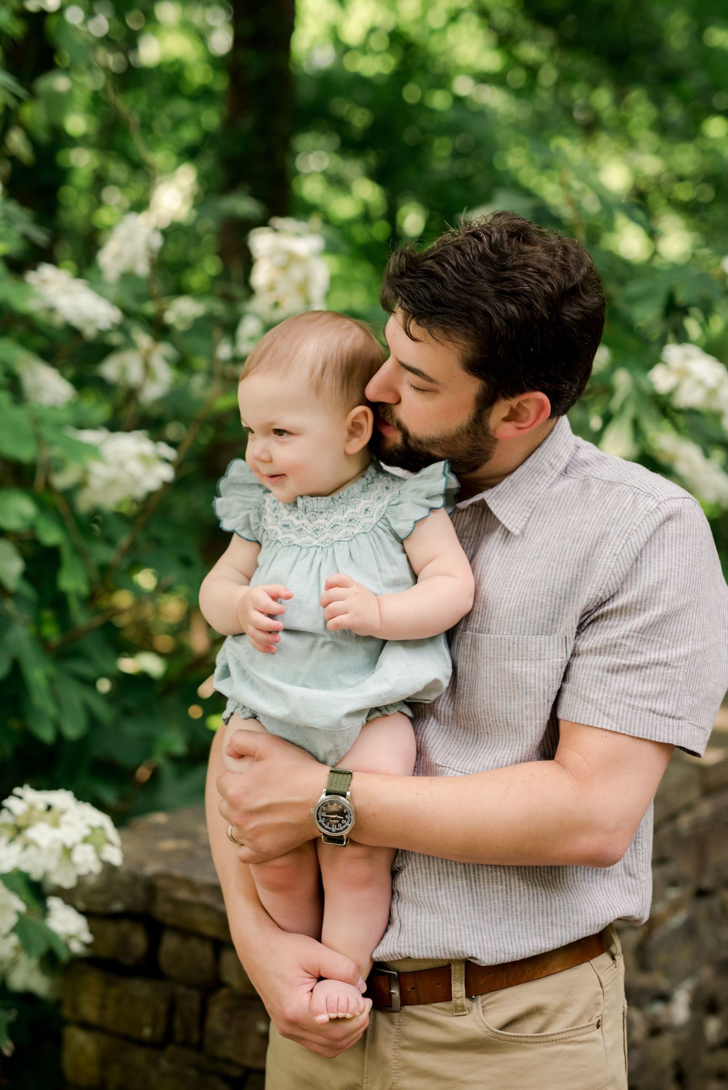 Adult holding and kissing a baby in a light blue dress against a green garden backdrop.