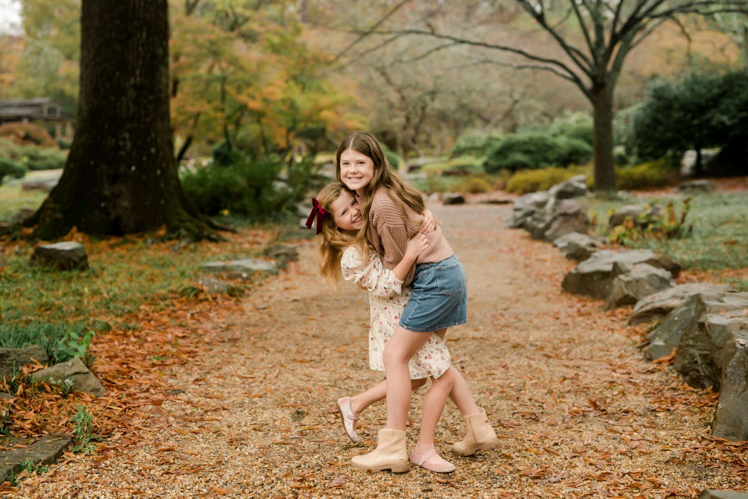 Two young girls hugging and laughing together on a fall garden path surrounded by autumn leaves.