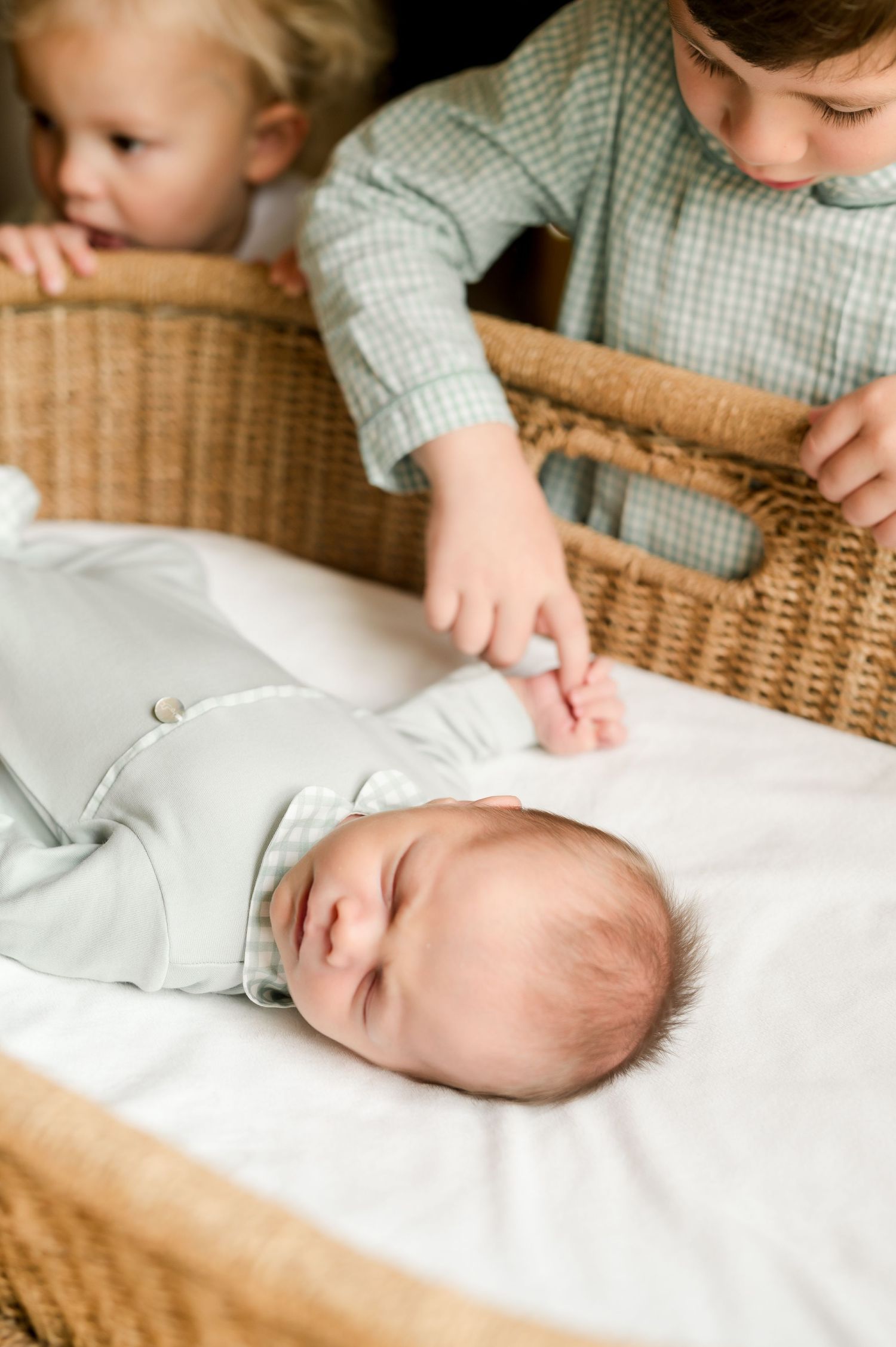 A sleeping newborn baby in a bassinet while two older siblings lean over to gently touch the infant's hand.