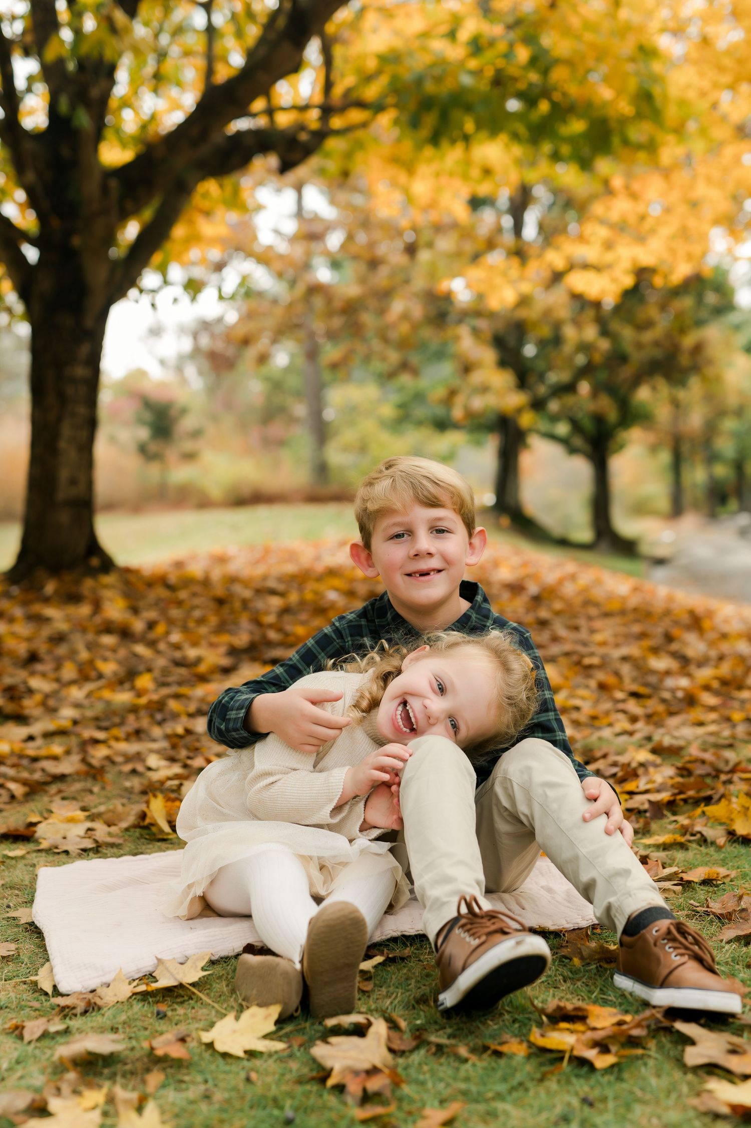 Two young siblings laugh and cuddle together on a blanket surrounded by golden fall leaves beneath autumn trees.