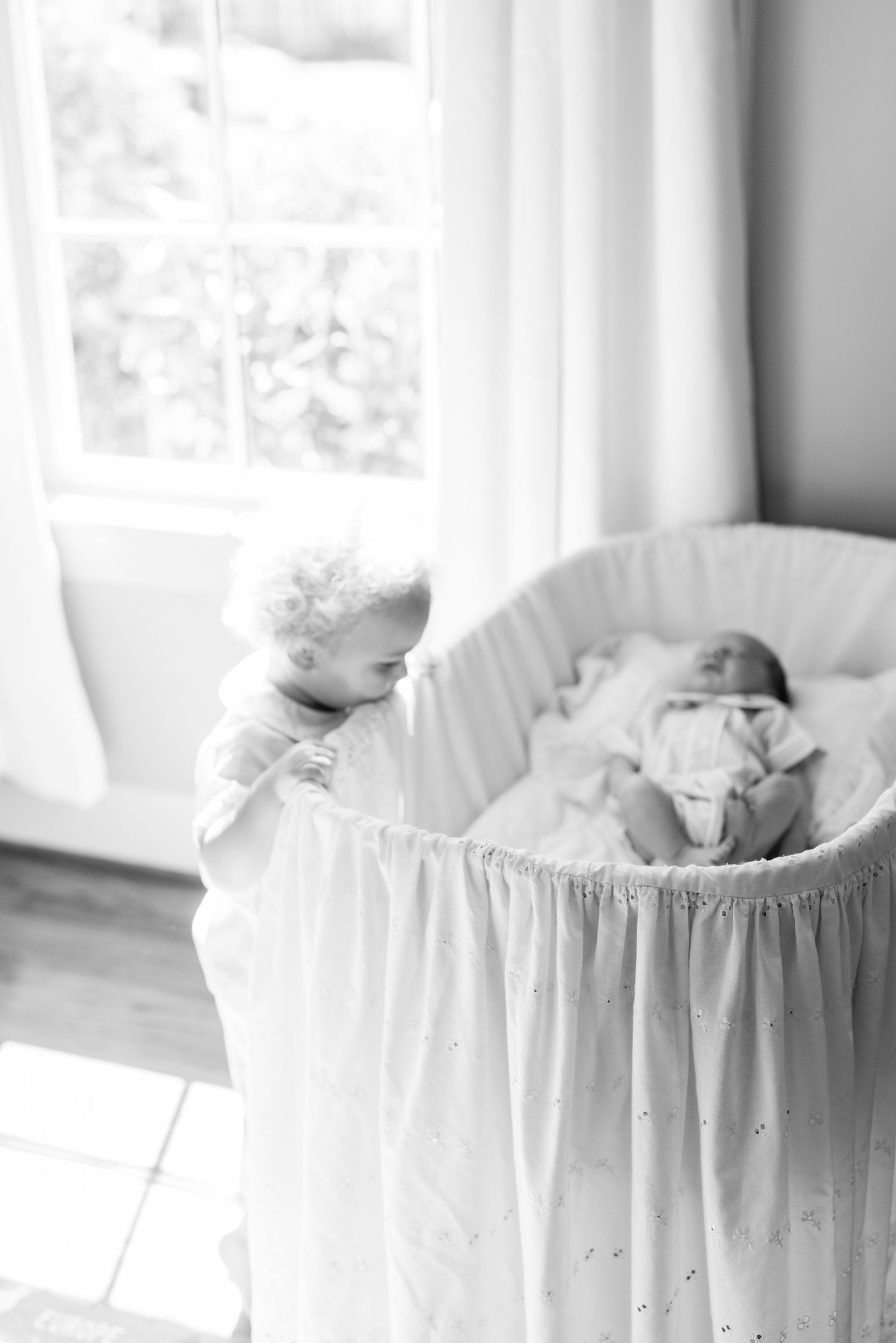 Black and white photo of a child peeking into a white bassinet near a window.