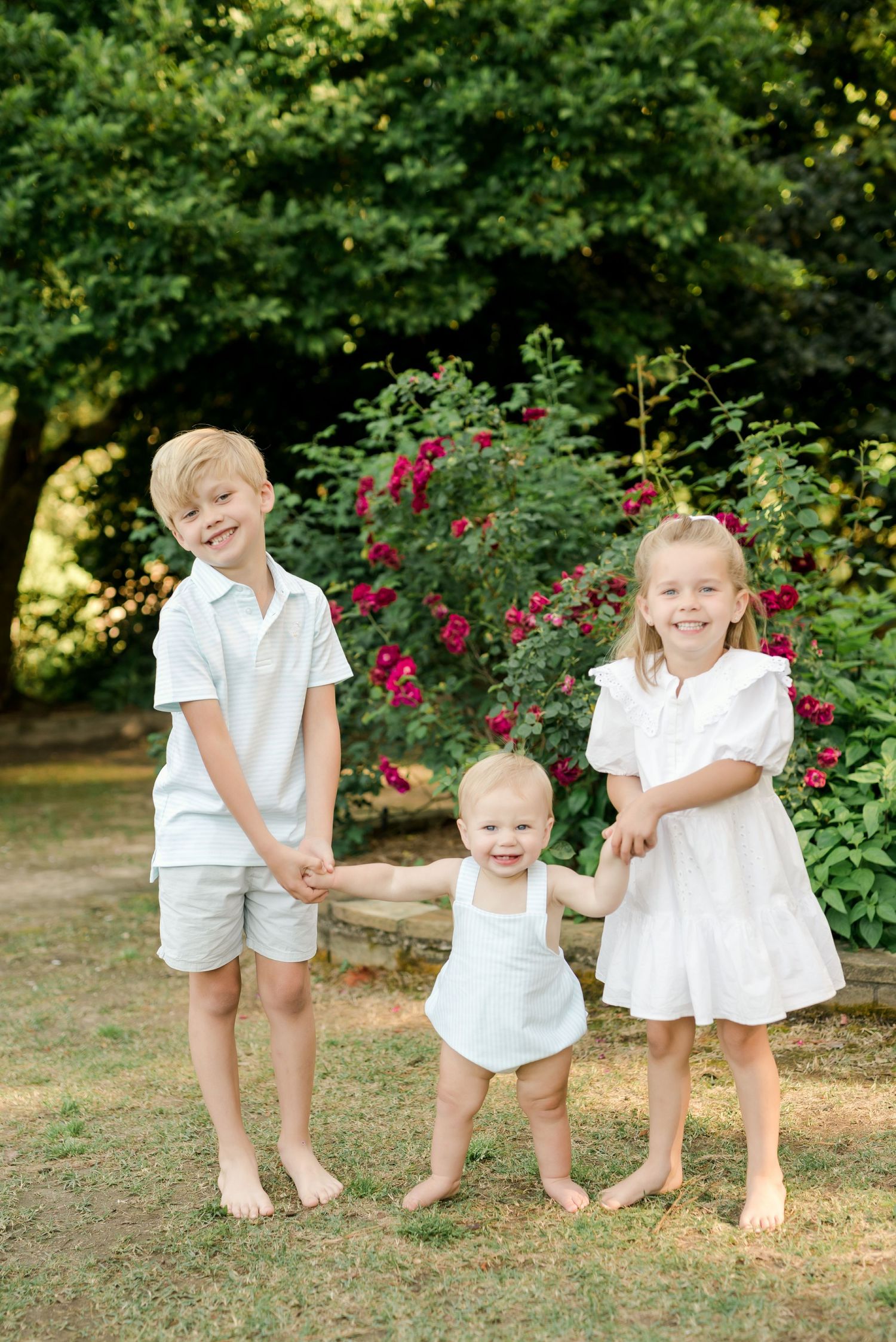 Children dressed in white outfits standing barefoot in a garden with blooming red roses in the background.