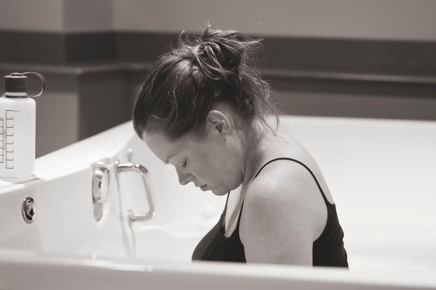 Black and white photo of person leaning over bathroom sink in contemplative pose.