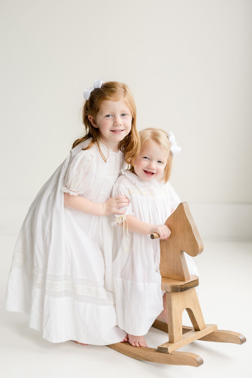 Two children in white dresses pose with a wooden rocking horse in a minimalist studio setting.