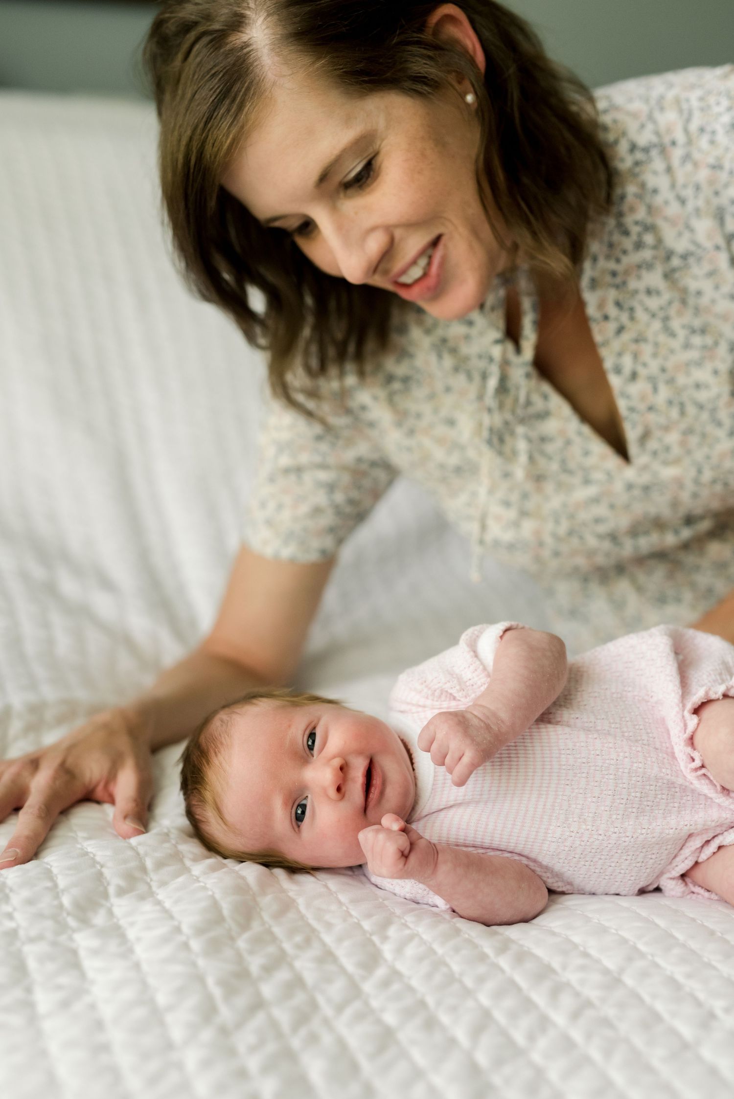 A baby in a pink outfit lies on a white quilted bed while being lovingly attended to.
