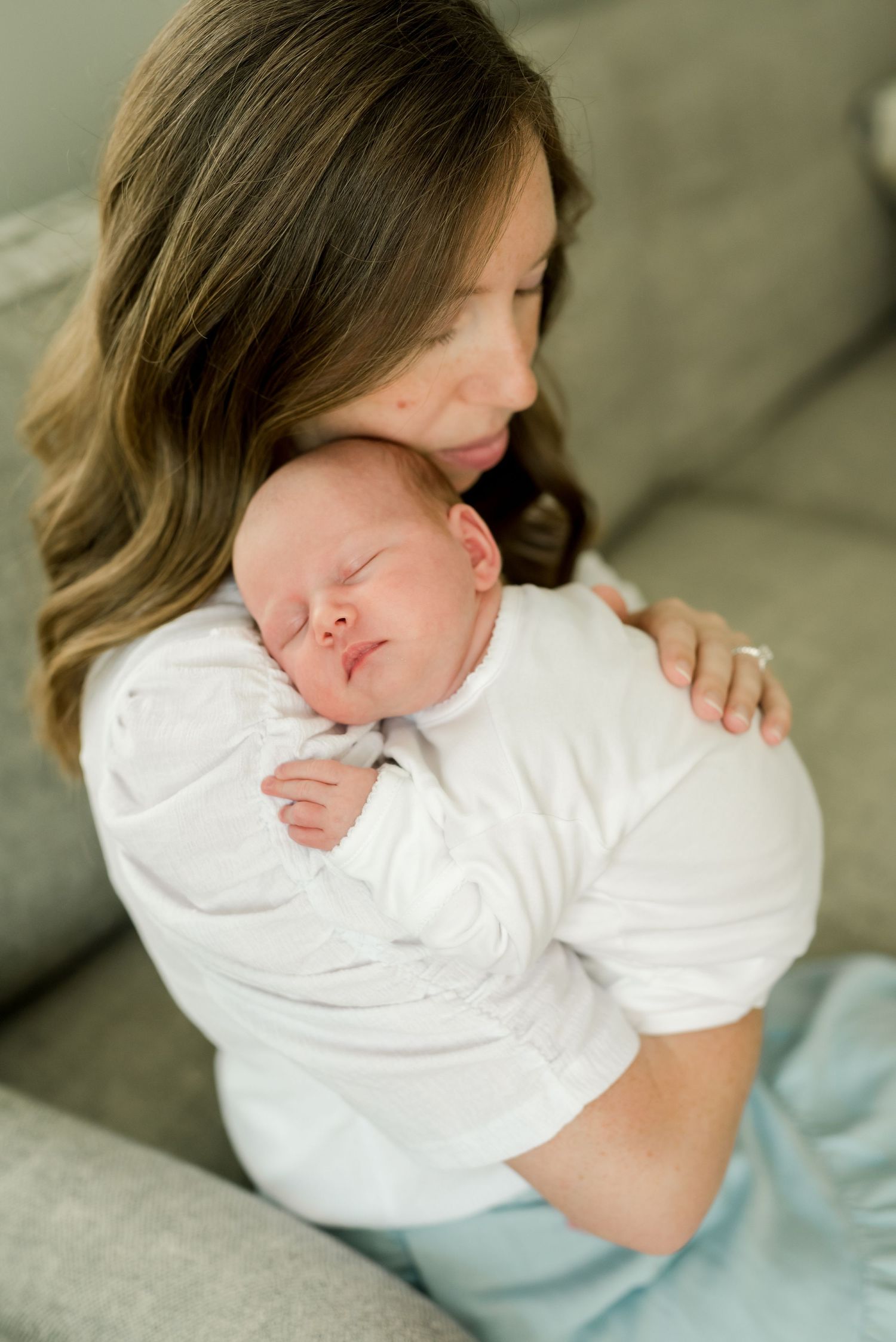 Tender moment between parent and sleeping newborn on grey sofa.