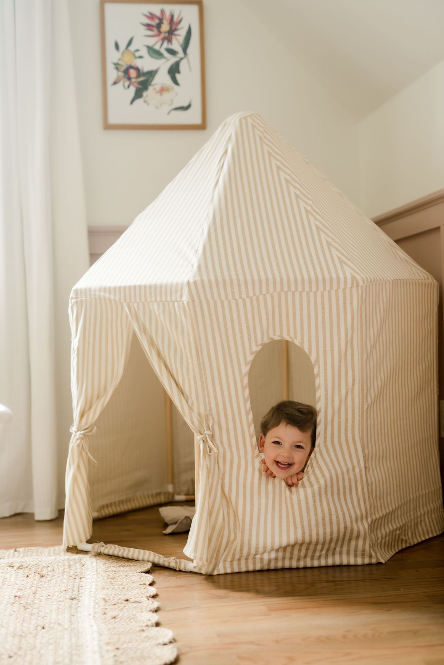 A striped cream-colored indoor play tent with window sits on hardwood floor beneath framed wall art.