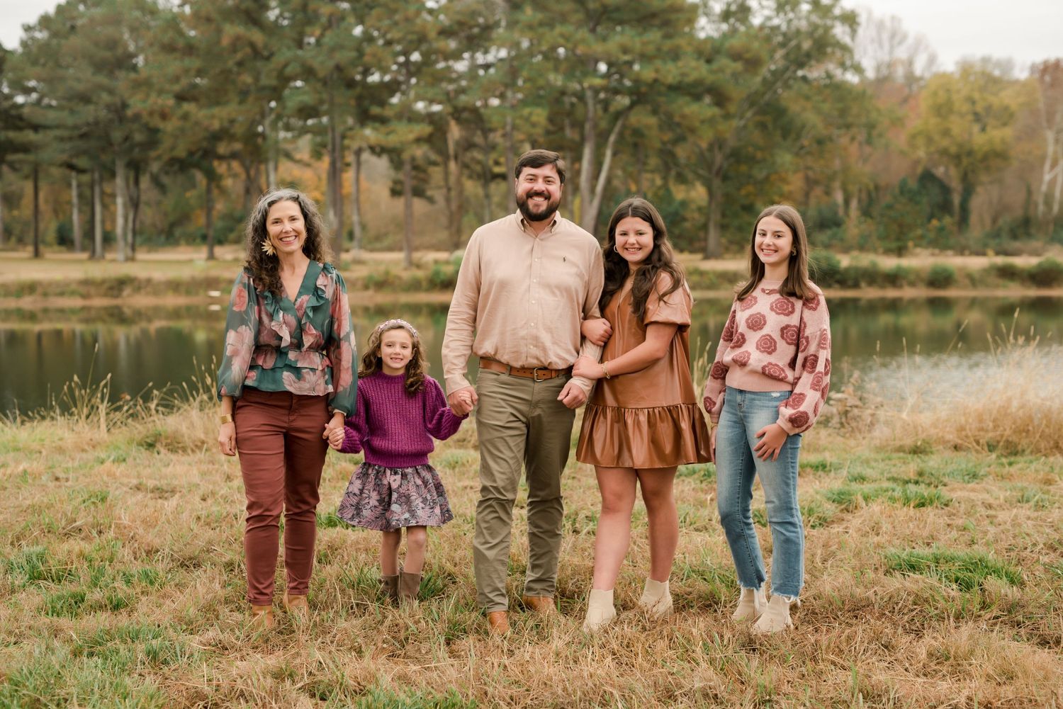 A family of five poses together on a grassy field with a serene lake and pine trees in the background.