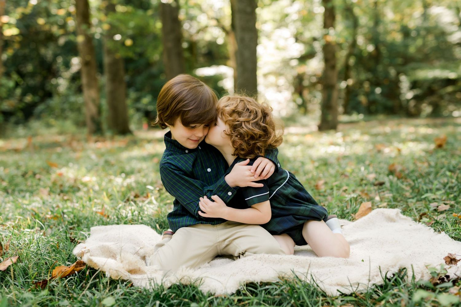 Two children embracing while sitting on a cream colored blanket in a forest setting.