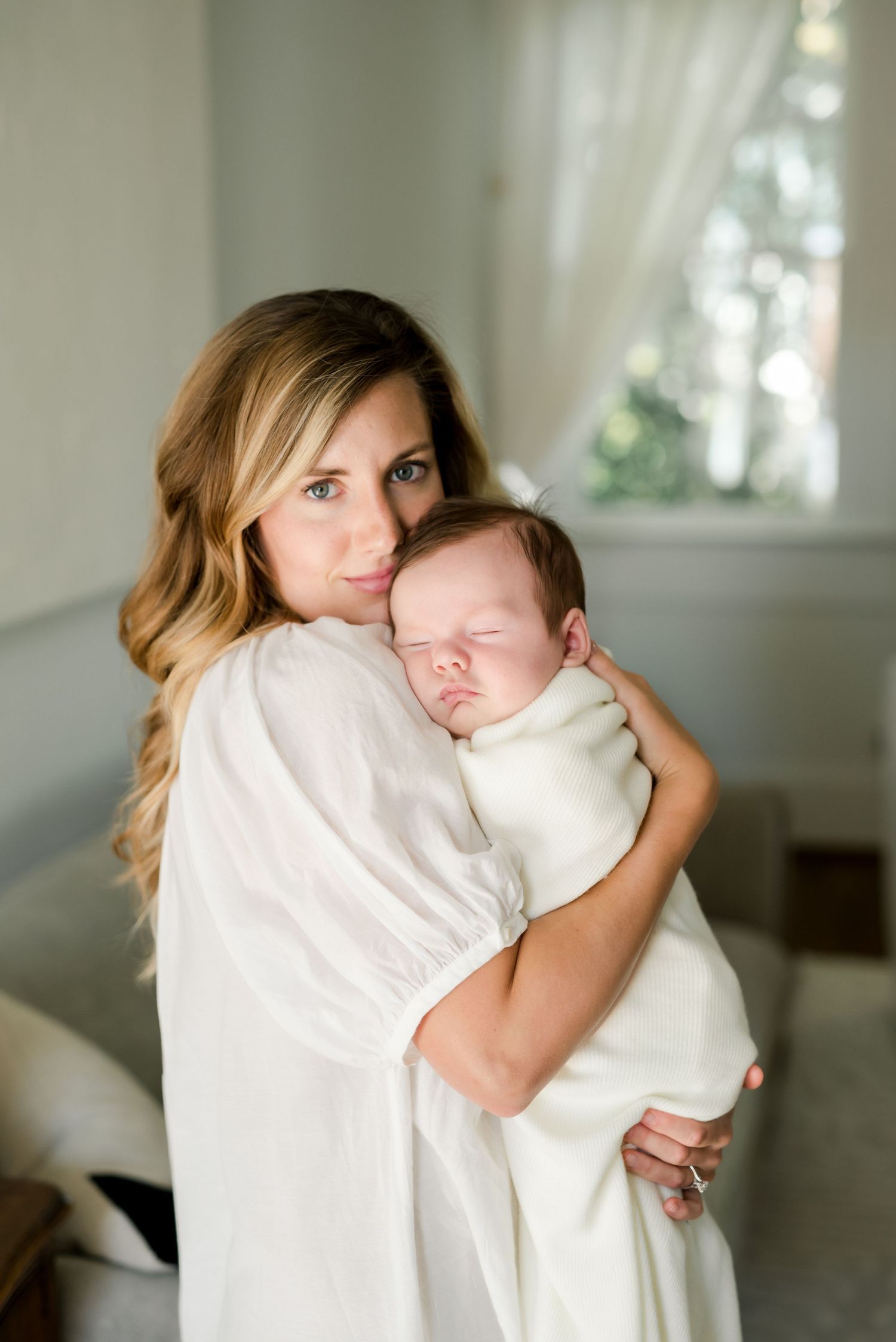 Tender moment between mother and sleeping newborn in white sunlit room.