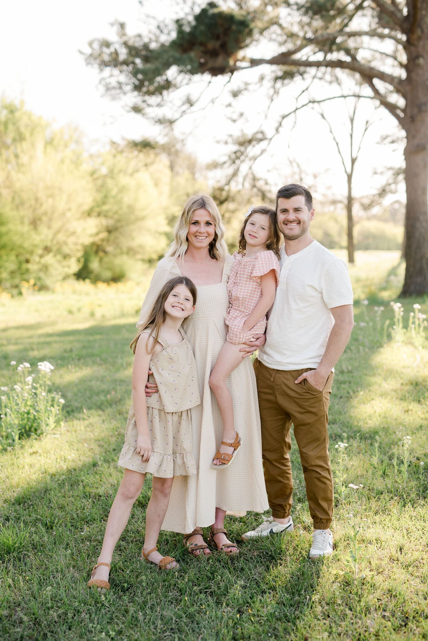 A family of four in coordinated neutral tones poses together in a sunlit meadow.