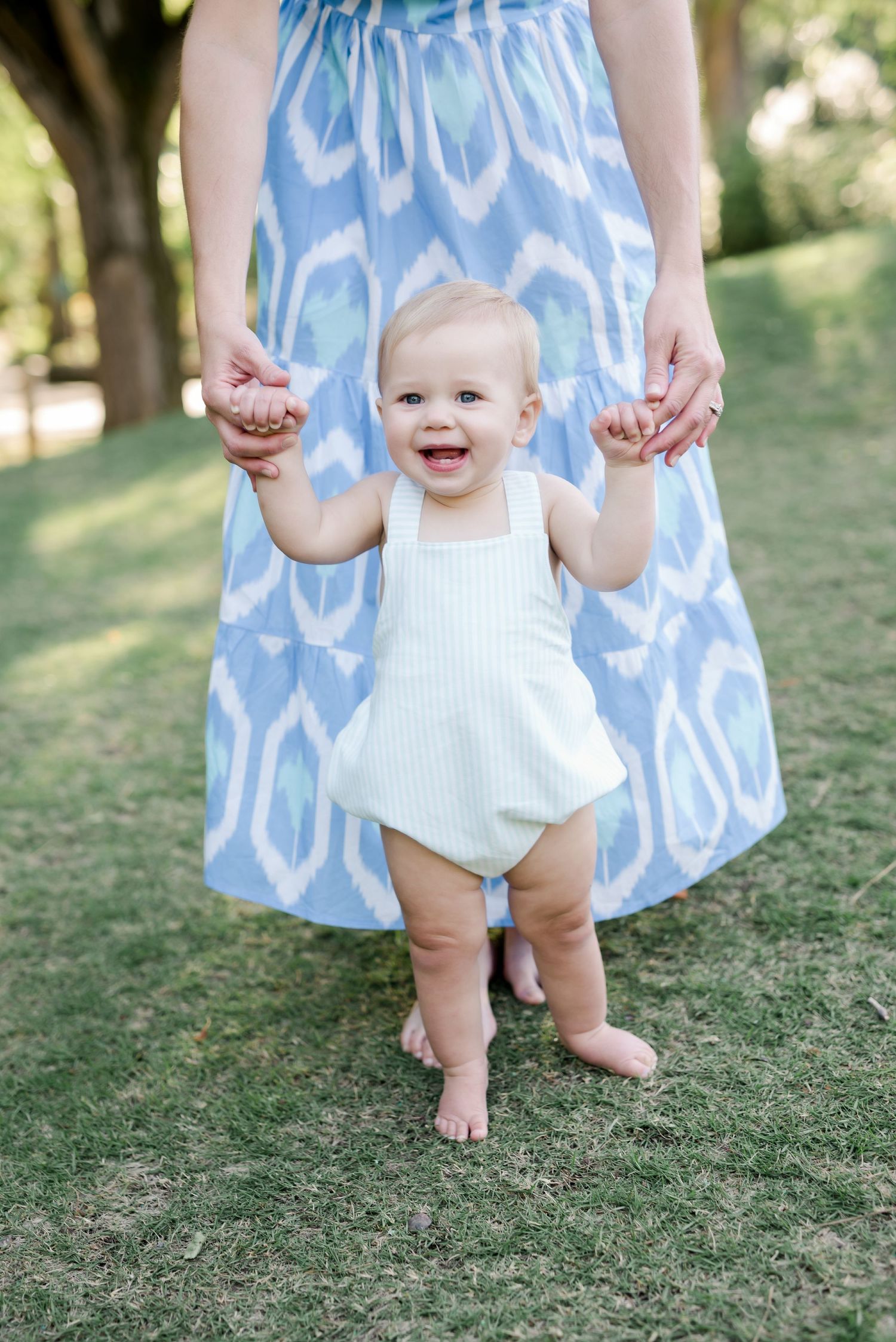 Small child in white outfit holding hands with adult wearing blue patterned dress outdoors.