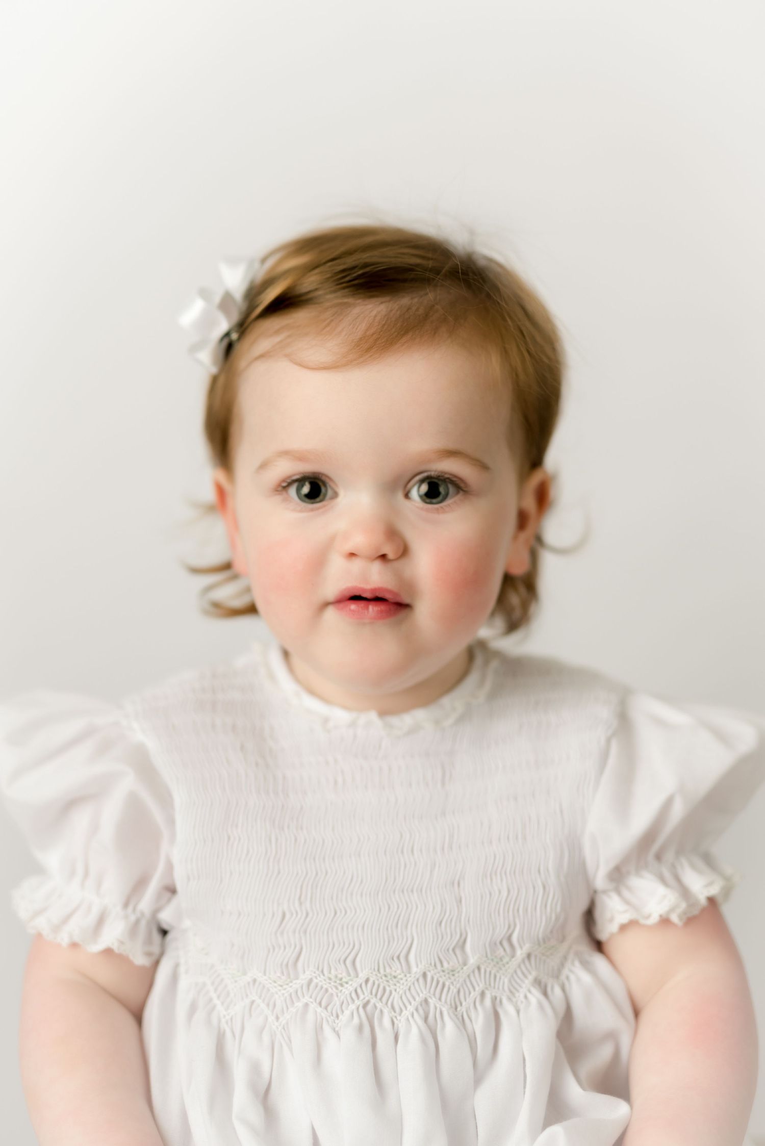Baby in white smocked dress with floral hair clip against white background.