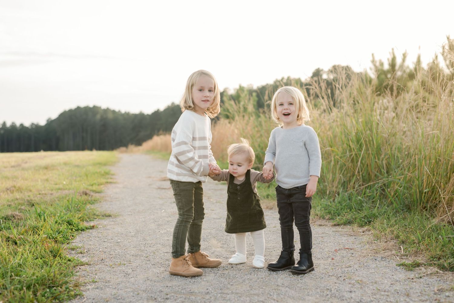 Three young children standing together on a dirt path with tall grass and trees behind them.