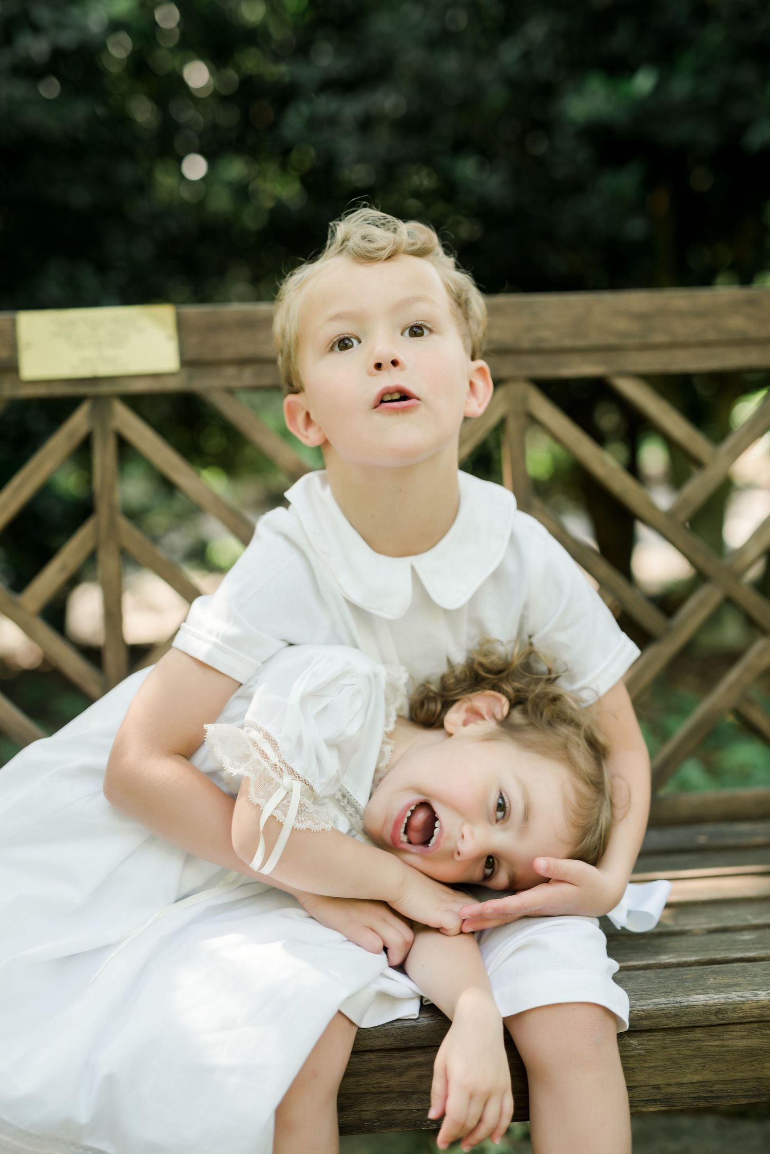 Playful siblings share lighthearted moment on wooden bench in garden setting.