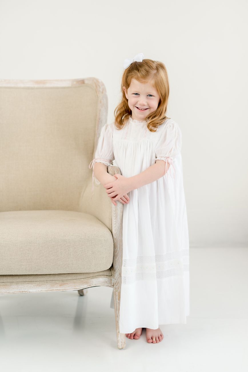A child in a white nightgown stands beside a beige couch in a bright minimalist setting.