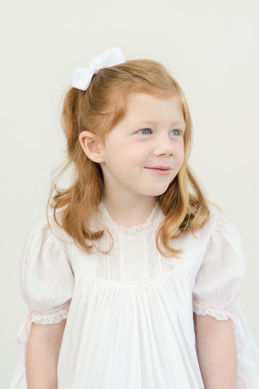 Young child in white dress with red hair and bow smiling against light background.