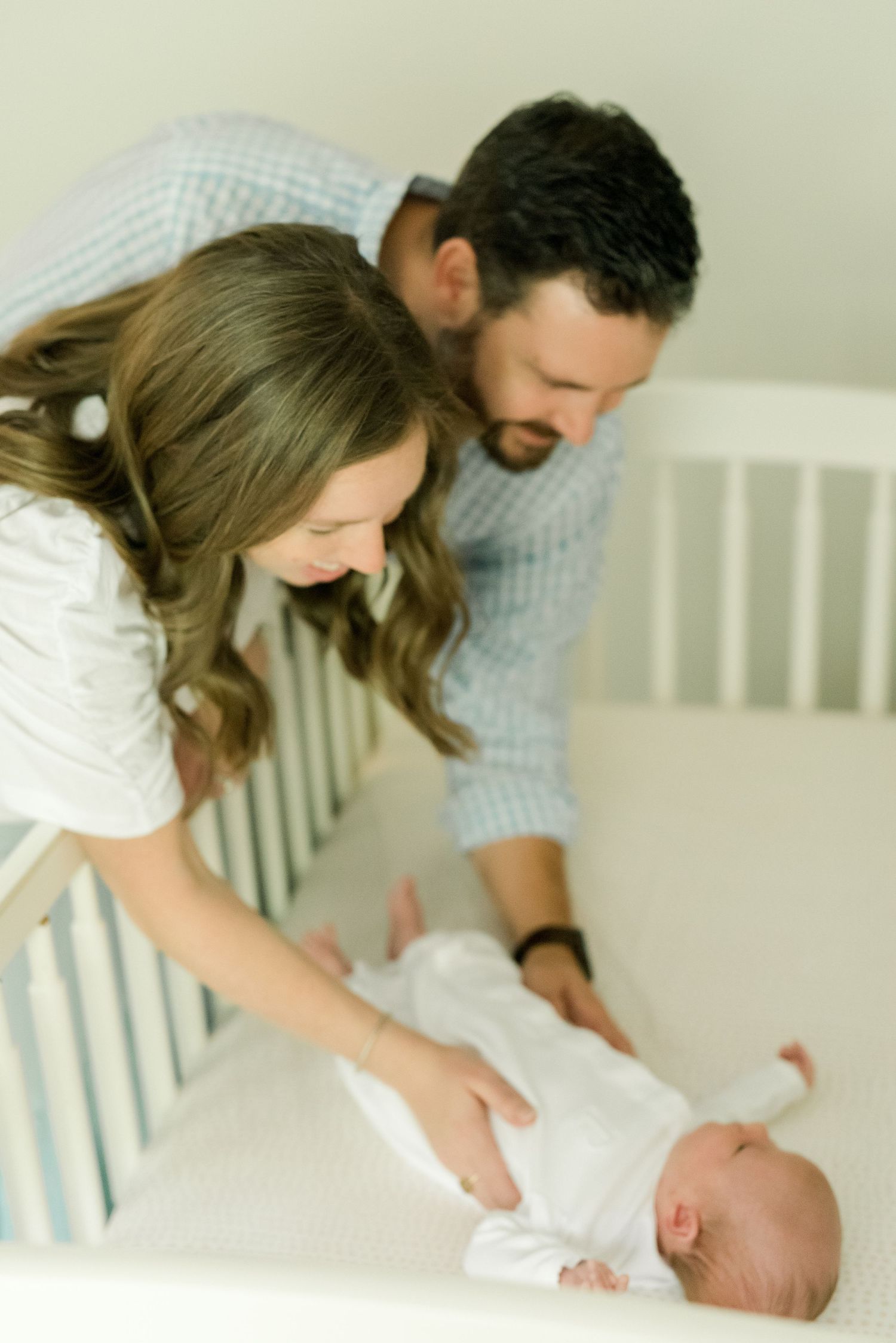 Parents lean over white crib looking down at their newborn baby.
