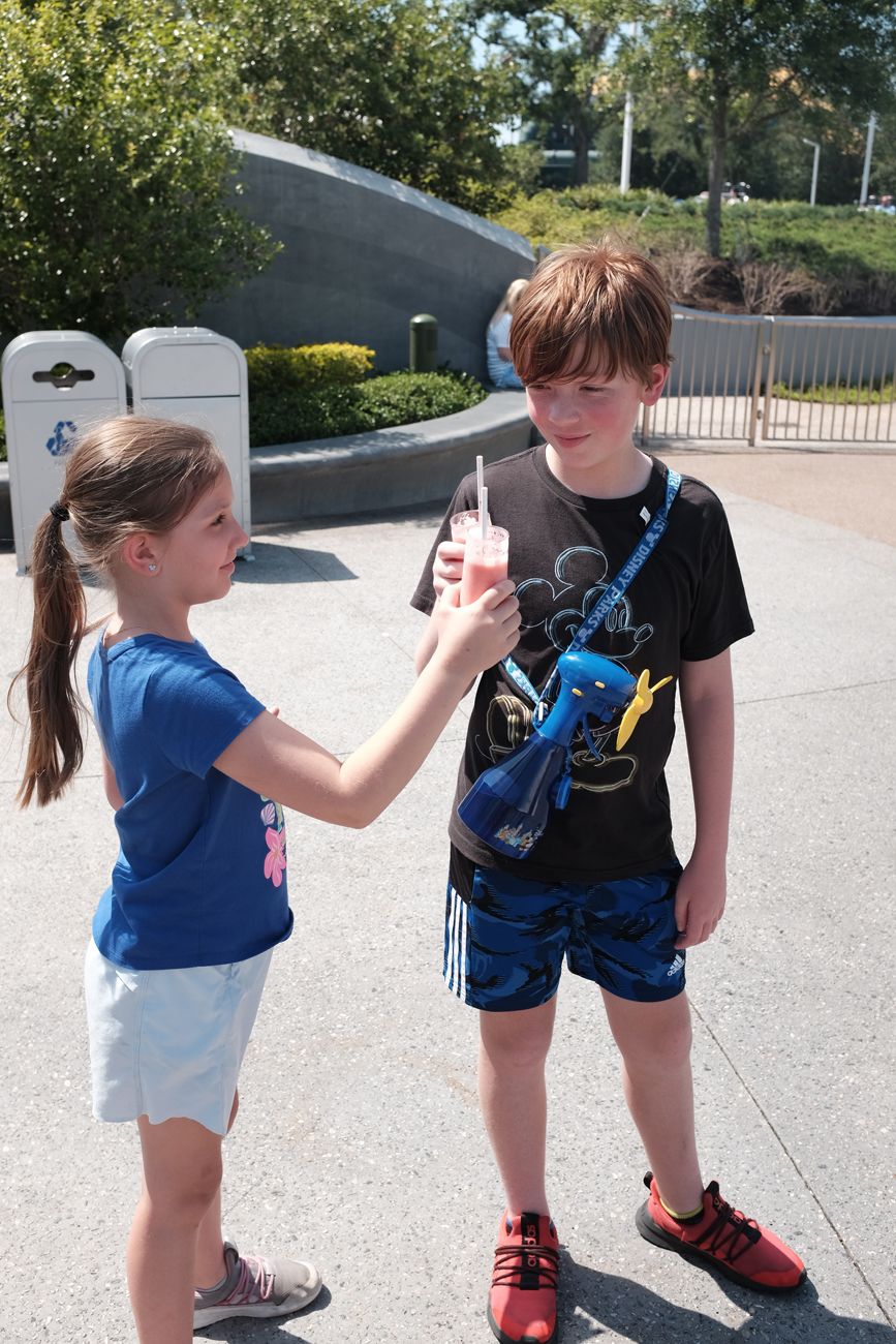 Two children share a drink together while standing outside in casual summer clothing.