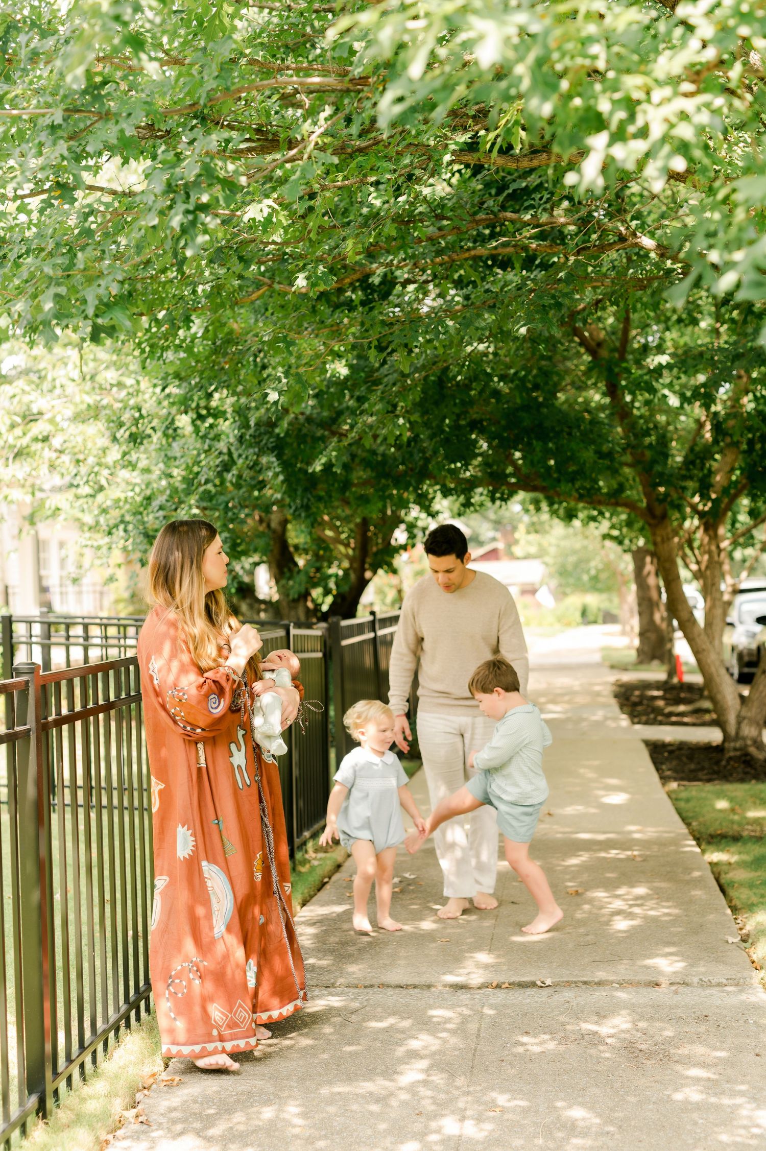 Family of four strolling on a tree-lined sidewalk, mother in rust floral dress holding newborn, two toddlers playing.