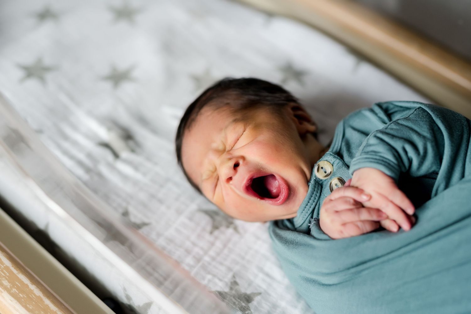 A newborn baby yawns while lying on star-patterned bedding in a hospital bassinet.