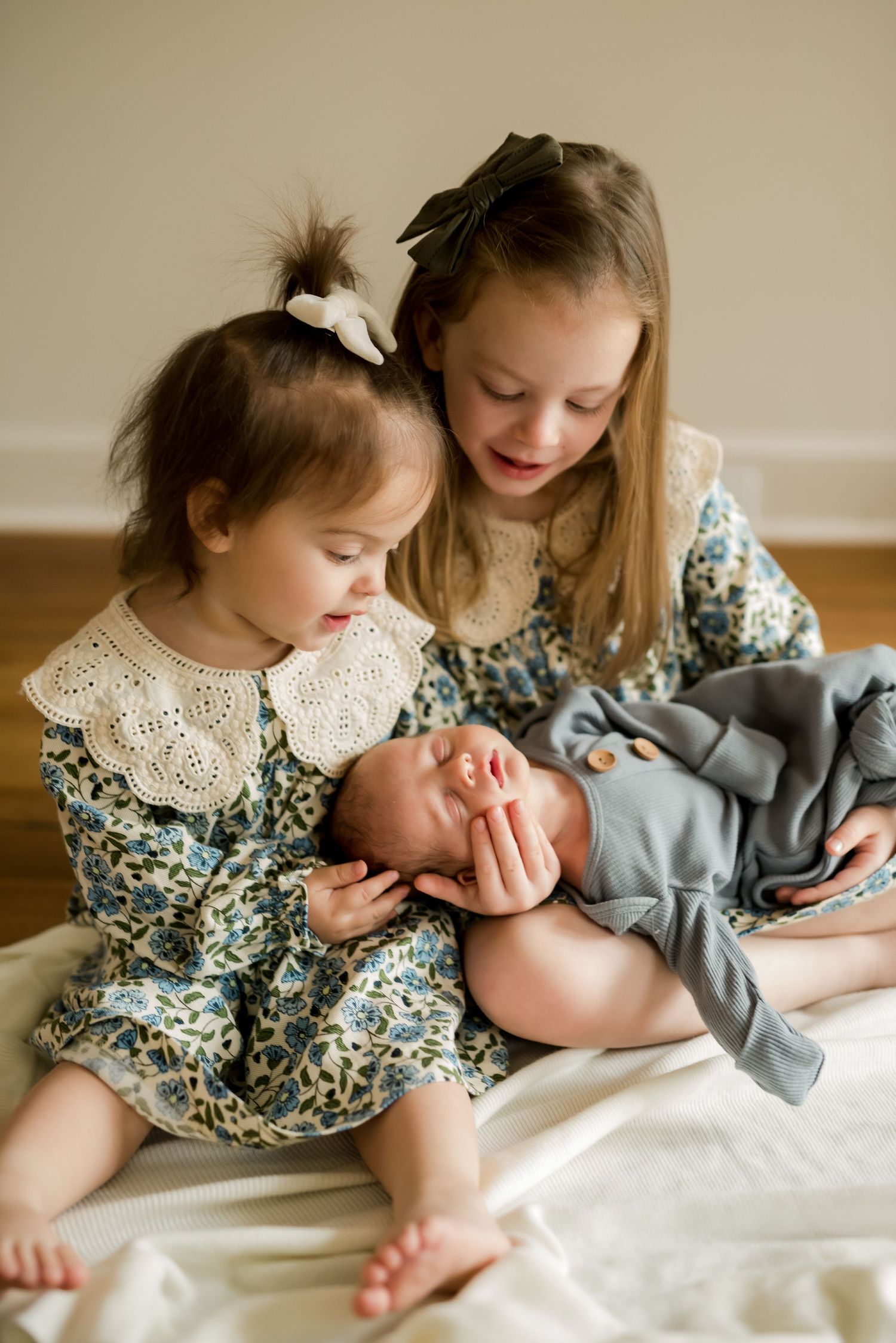 Two young children in matching floral dresses tenderly holding sleeping baby on white bedding.