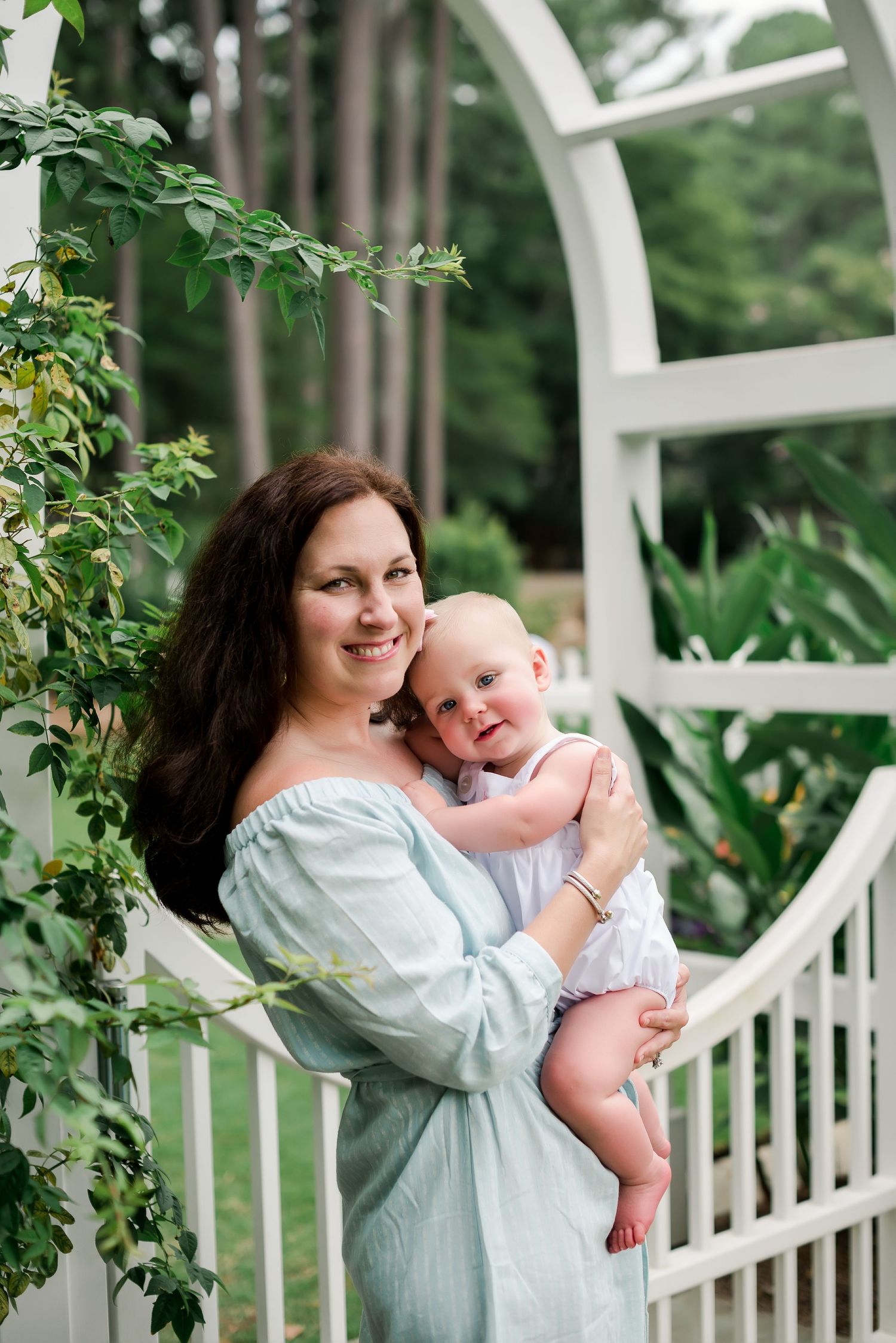 Mother and baby share an intimate moment by a white garden trellis surrounded by greenery.