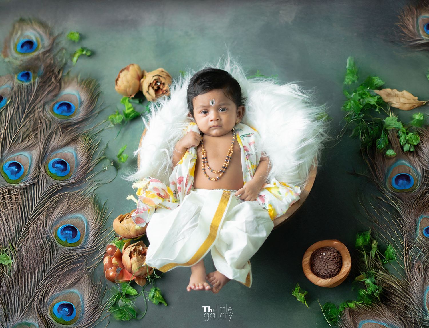 Baby in traditional Indian attire lying on white cushion with peacock feathers and floral decorations.