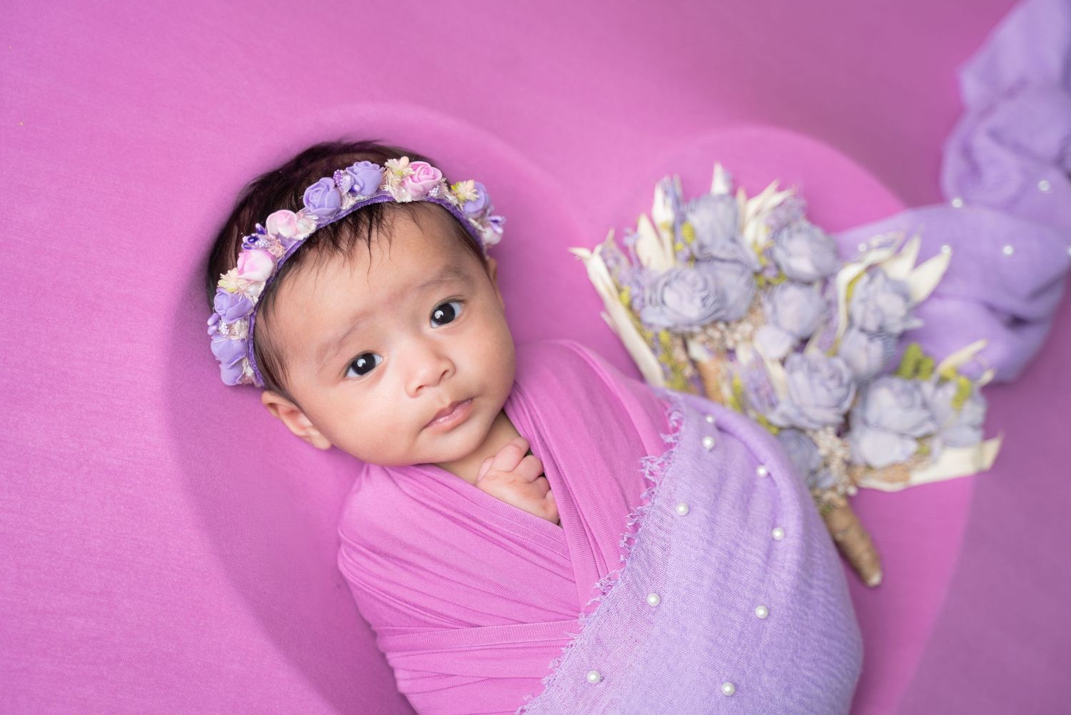 Dreamy newborn portrait with purple and pink tones featuring floral decorations.