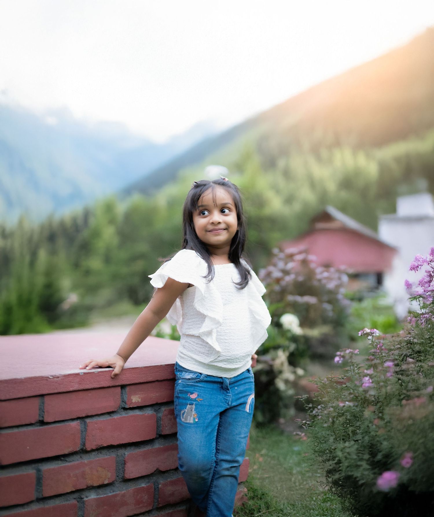 Child in white top and jeans leans on brick wall with mountain landscape behind.