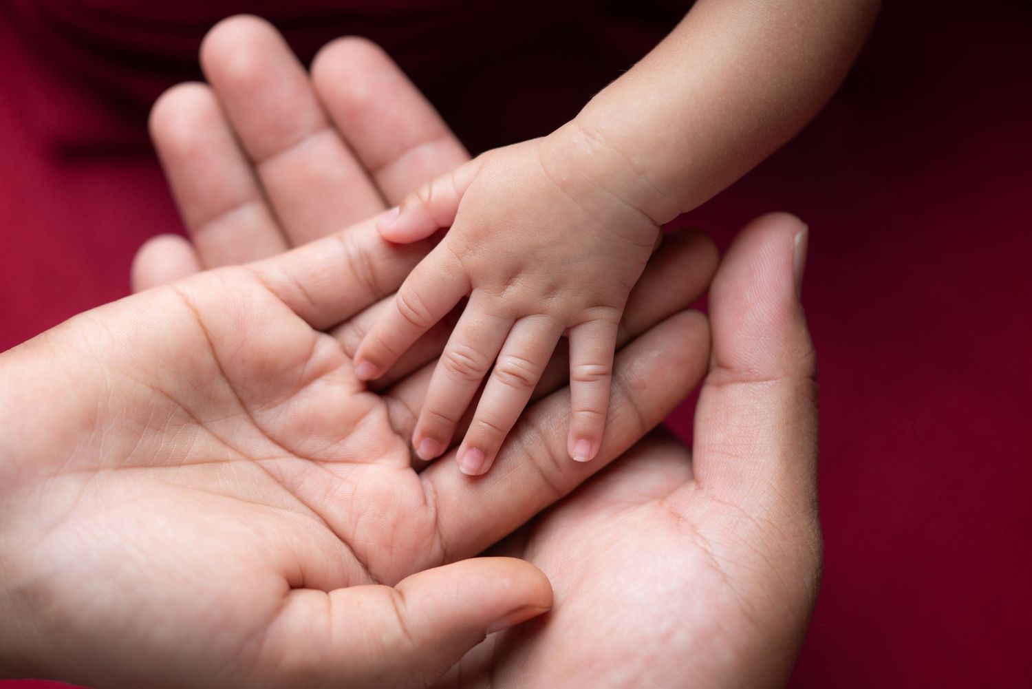 Close-up series of gentle hands holding a newborn baby against a deep red background.
