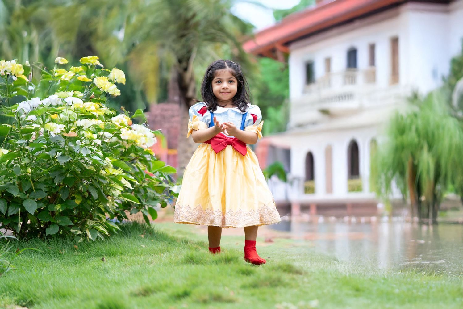 Child in yellow dress with red boots stands in garden with blooming flowers.