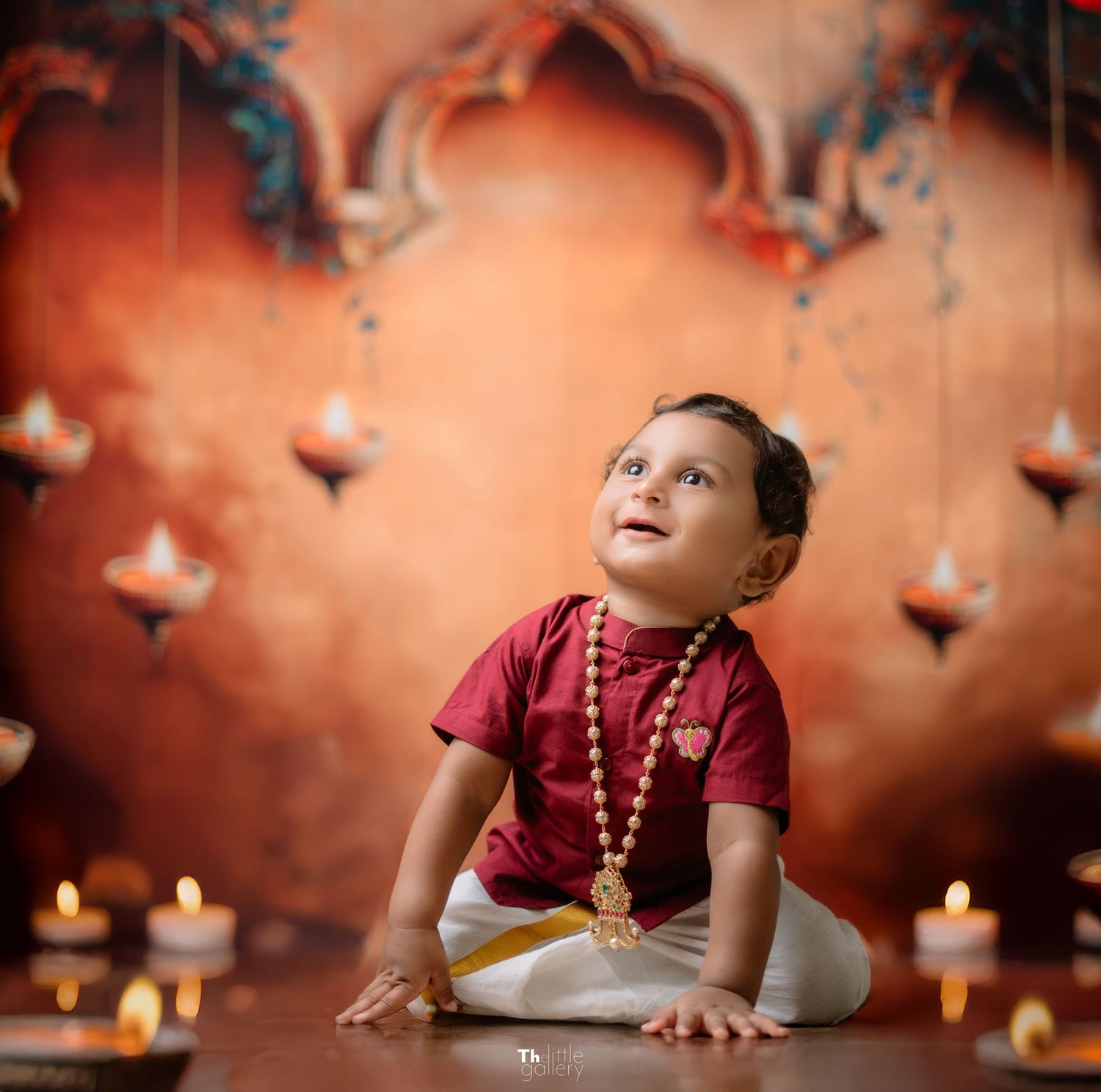 Child in red shirt sits among lit candles and traditional decor in warm-toned setting.
