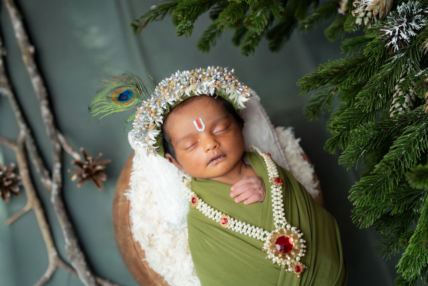 Sleeping newborn in green wrap with crystal crown and peacock feather decoration.