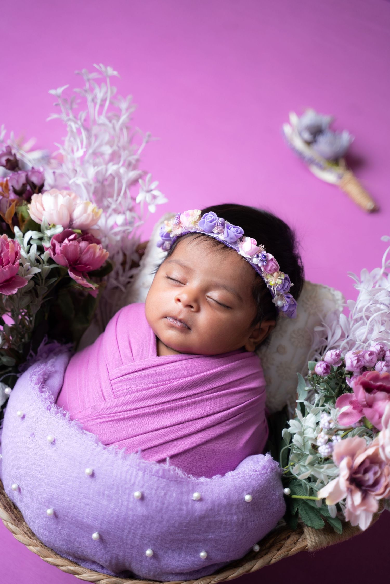 Peaceful newborn portrait on purple cushion surrounded by delicate flowers and soft pink wrapping.