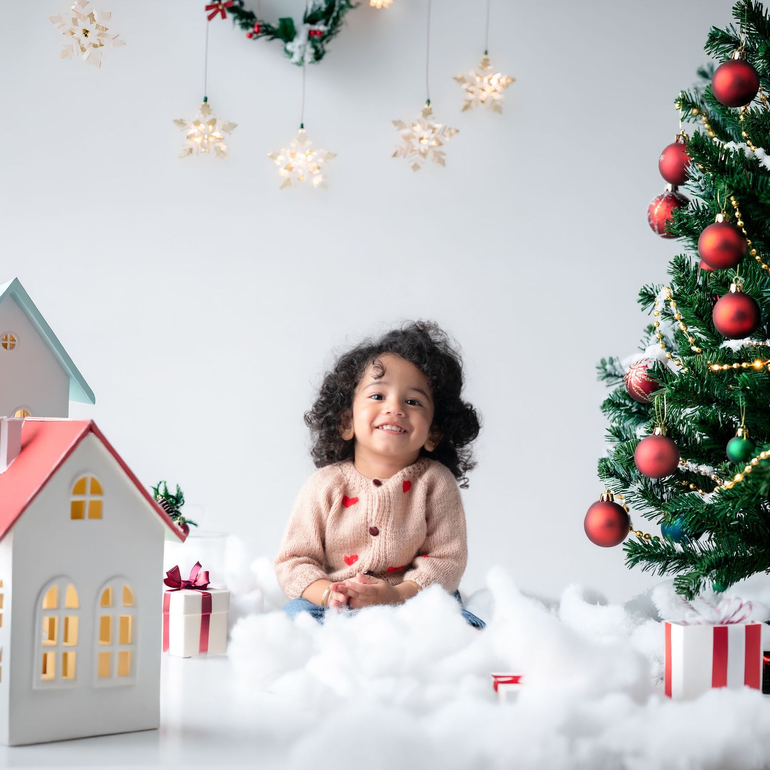 Child in pink sweater sits among Christmas decorations with toy house and tree.