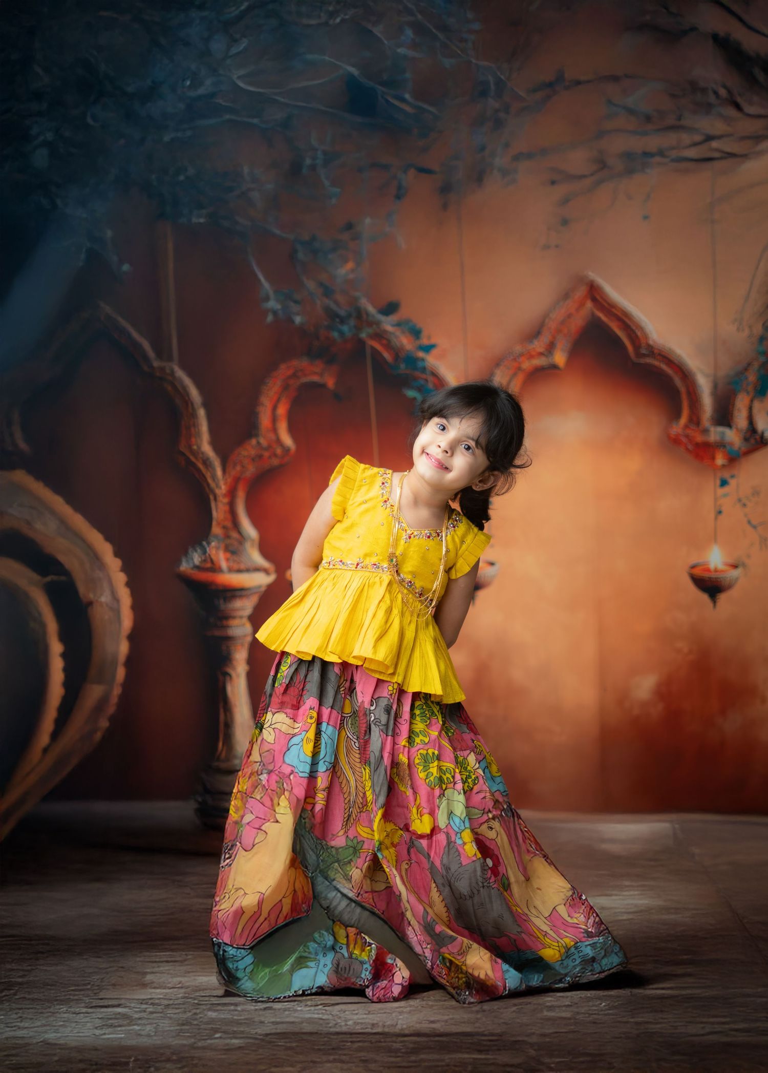 Child wearing vibrant yellow and floral dress poses playfully against ornate orange backdrop.
