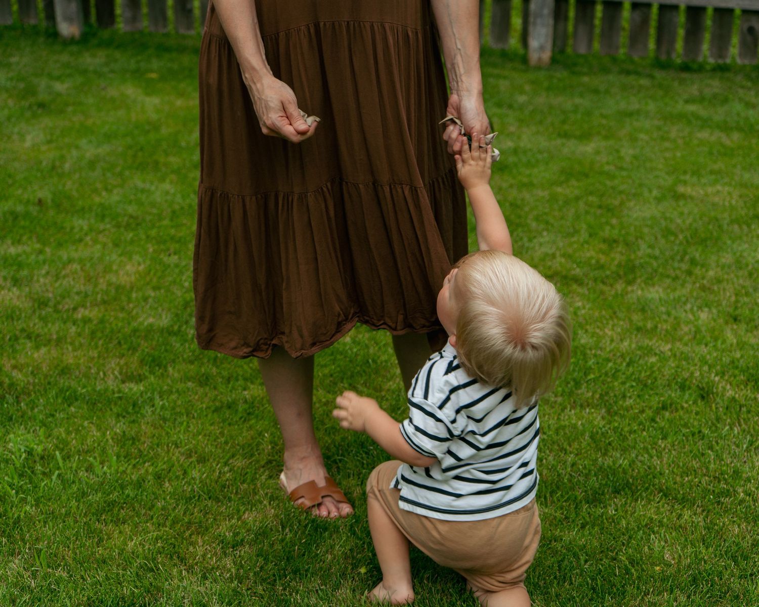 A toddler in a striped shirt holds their mother's hand while learning to walk on green grass outdoors.