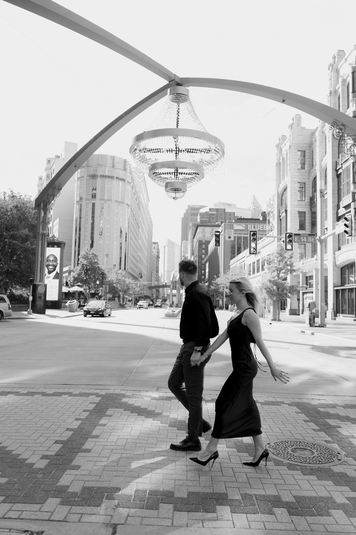 Black and white photo of a couple holding hands walking on a city sidewalk under an elegant chandelier arch.