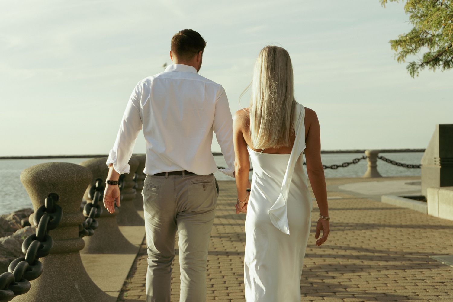 Couple walking hand in hand along a waterfront promenade, man in white shirt and woman in elegant white dress.