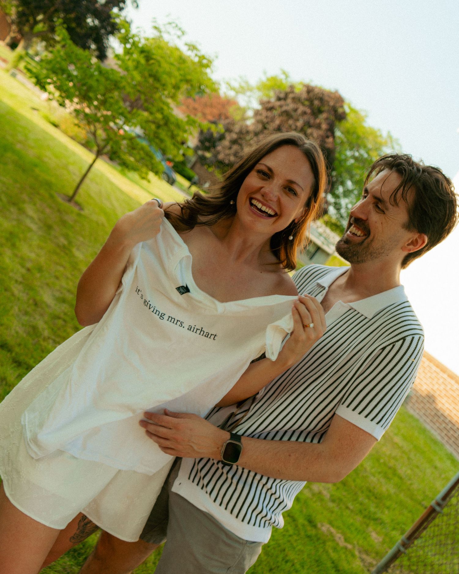 Happy couple laughing outdoors in a sunny park, woman holding a white shirt reading future mrs. airhart.