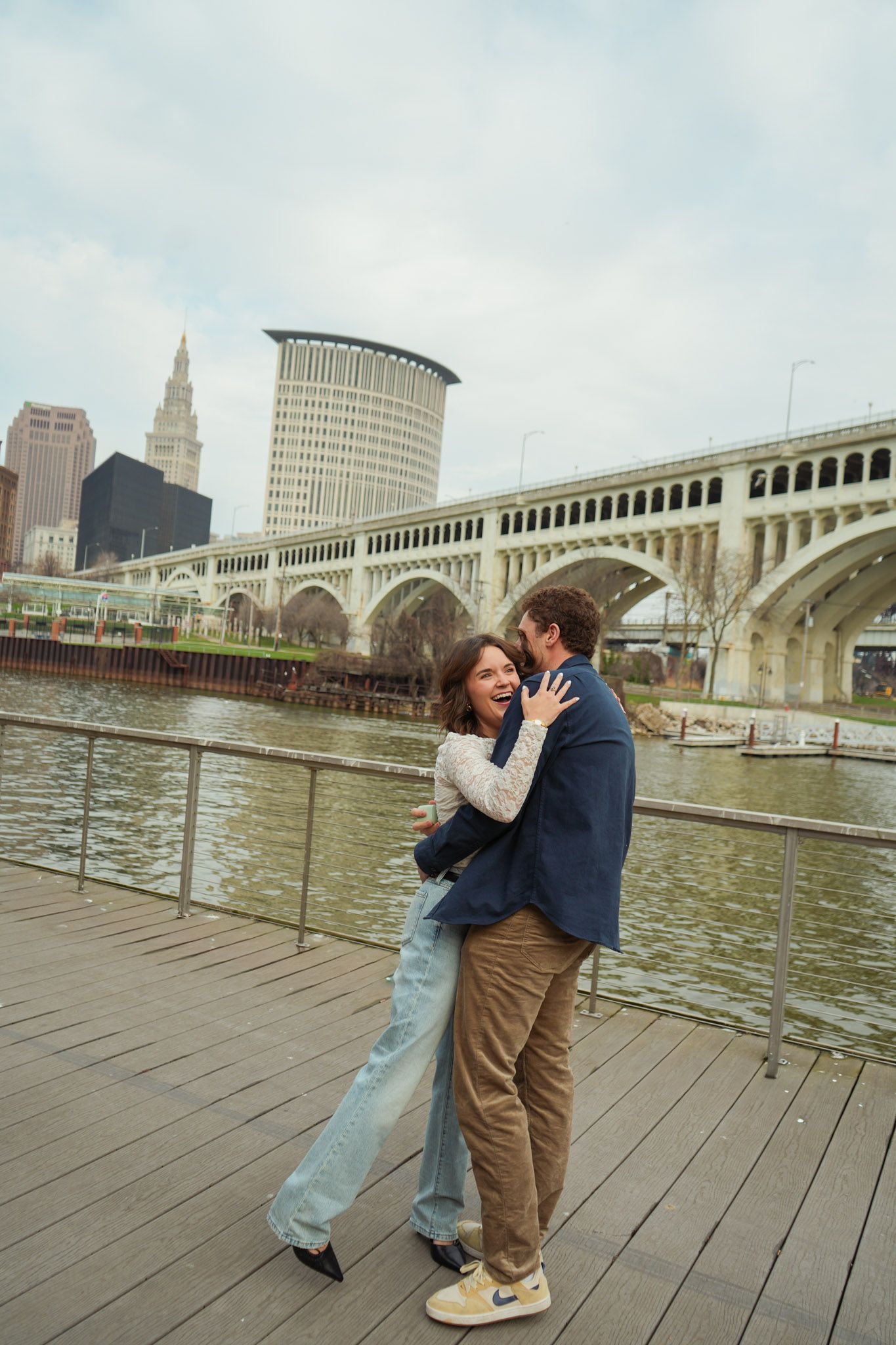 A couple shares a romantic moment on a wooden pier with Cleveland's iconic bridge and skyline in the background.
