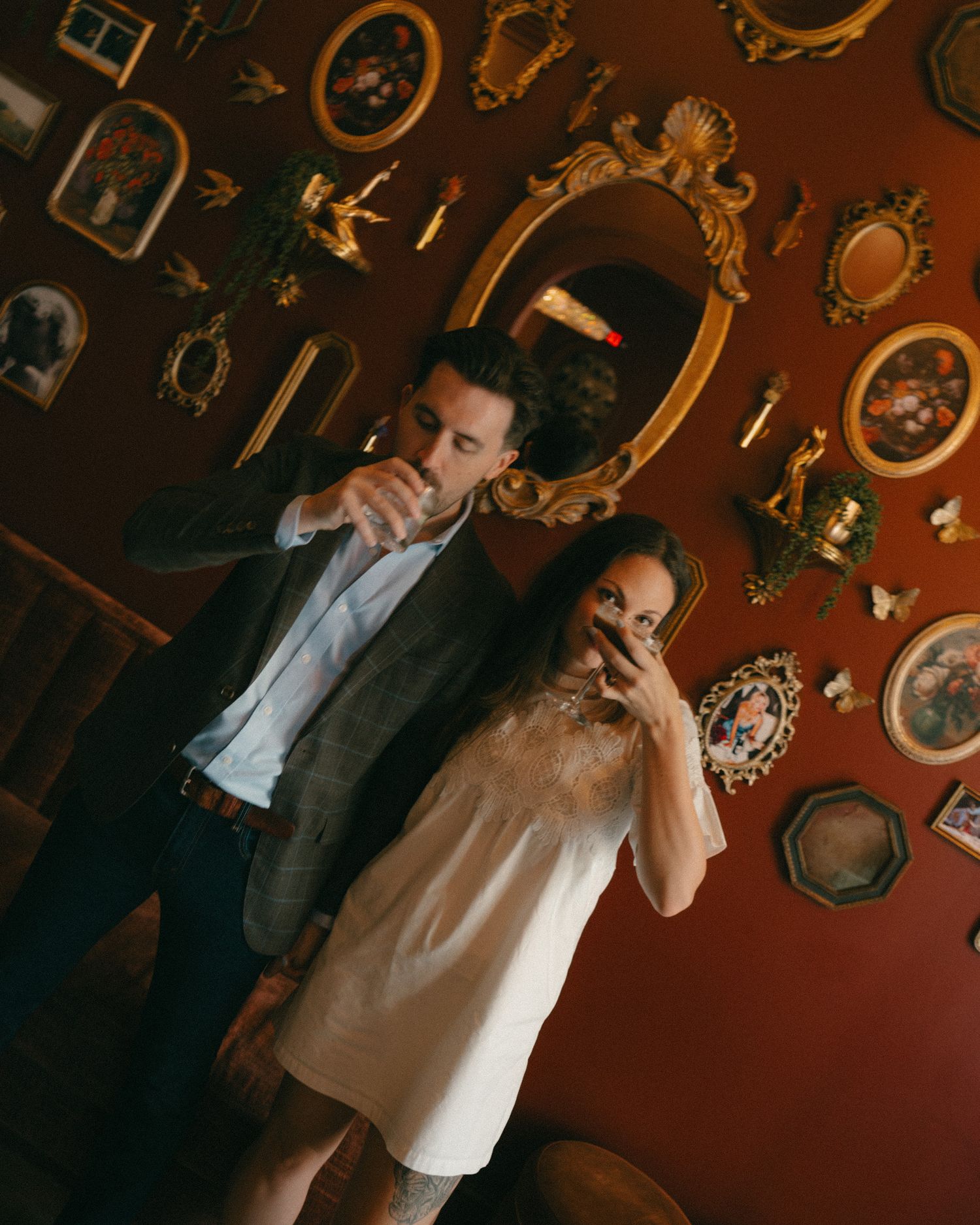 Couple posing in front of ornate gold-framed artwork on a red wall, woman in white dress, man in dark suit.